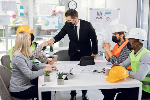 Group of five multiethnic colleagues wearing medical mask while sitting at table and fist bumping. Business meeting during coronavirus pandemy. Concept construction industry.
